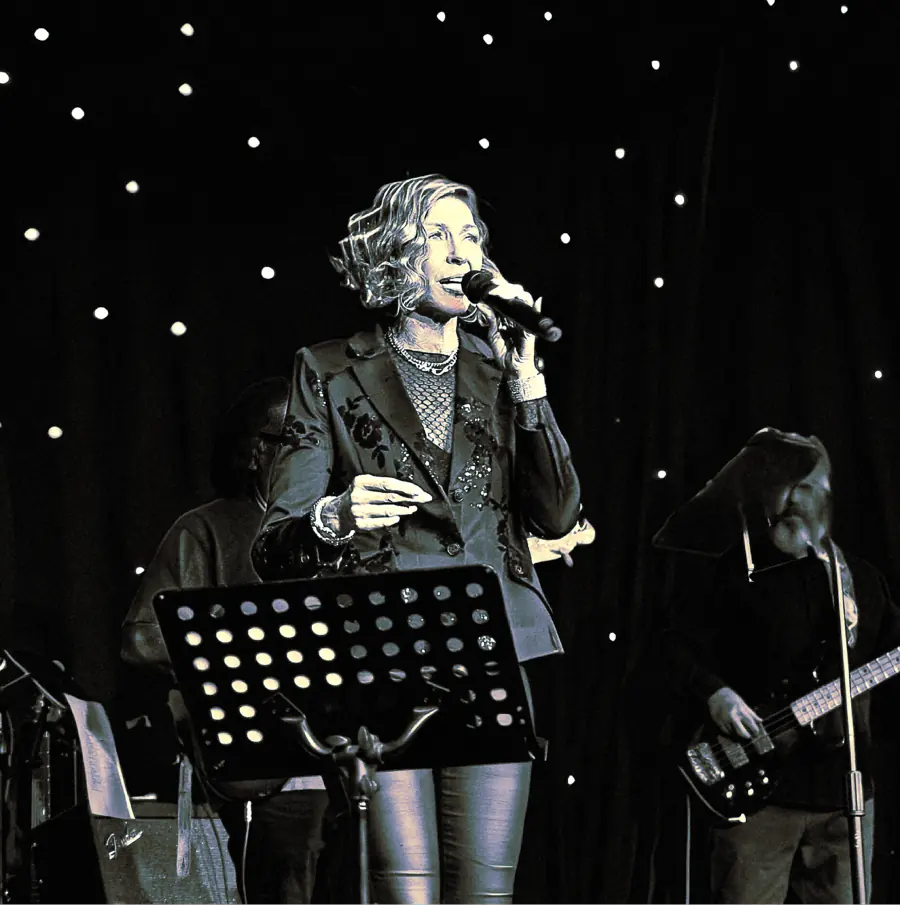 Black and white photo of Denise Choate singing on stage, musicians behind her under small lights