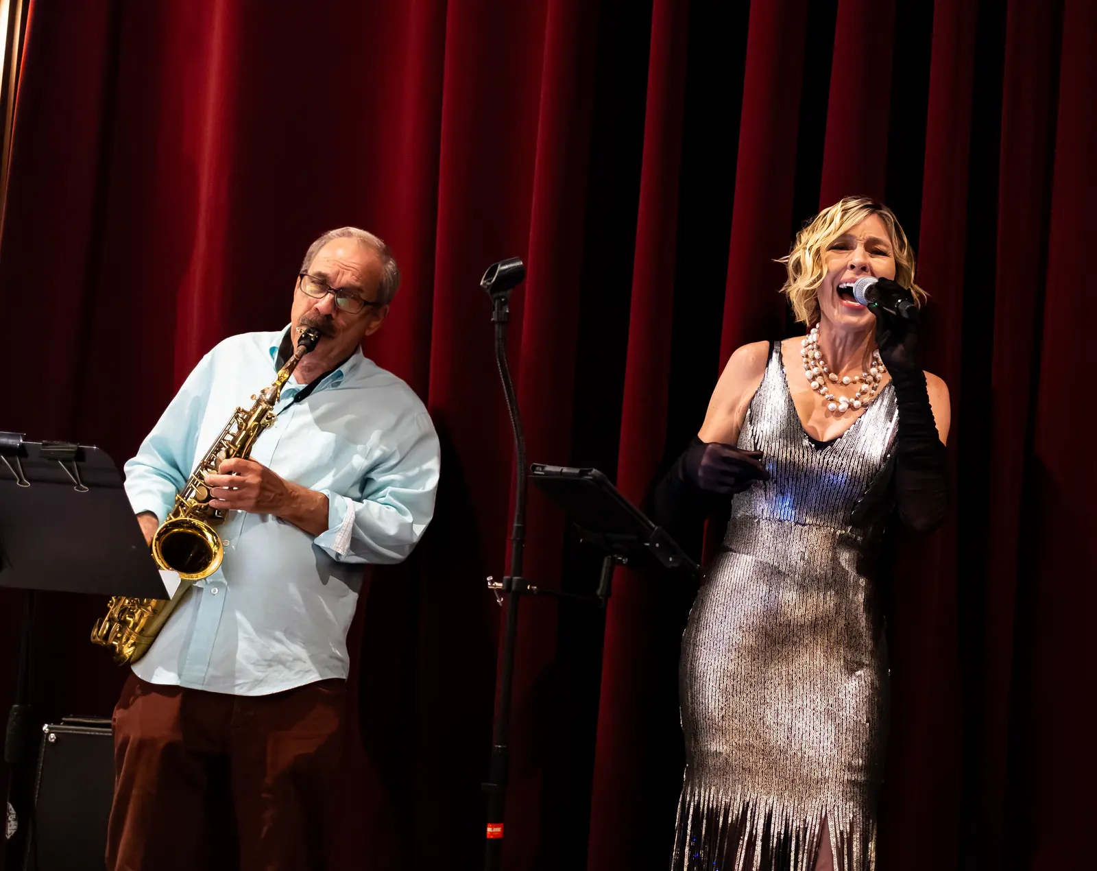 Denise Choate singing into a microphone beside saxophonist Paul Contos, in front of deep red stage curtains